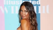 Alex Aust poses in front of a multi-colored pastel background and smiles over her shoulder at the camera.