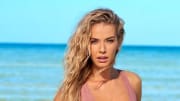 Olivia Jordan poses before the bright blue ocean in a light pink knit one-piece with her blonde hair in a light beach wave.