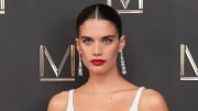 Sara Sampaio poses in a white dress and a red lip in front of a gray backdrop.