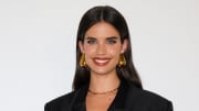Sara Sampaio poses in front of a white backdrop in a black blazer, red lip and oversized gold earrings.