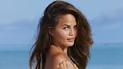 Chrissy Teigen poses in front of the ocean and looks back over her shoulder at the camera.
