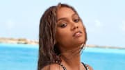 Tyra Banks poses in front of the bright blue ocean in a black and silver one-piece swimsuit and wet hair.