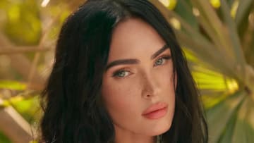 Megan Fox poses for the camera in front of vibrant green palm trees.