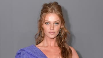 Cintia Dicker poses in a one-shoulder purple dress and wears her red hair in a half updo.