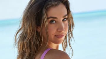 Katie Austin poses before the bright blue ocean in a pink one-piece and smiles softly over her shoulder.