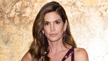 Cindy Crawford poses in front of a gold metallic backdrop. She wears oversized drop earrings and sports her brown hair in an oversized curl.