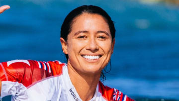 Malia Manuel sits atop a surfboard in the water and smiles for the camera.