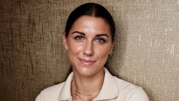 Alex Morgan poses before a textured gold backdrop in a cream-colored sweater and gold necklaces.
