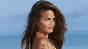 Chrissy Teigen poses in front of the ocean and looks back over her shoulder at the camera.