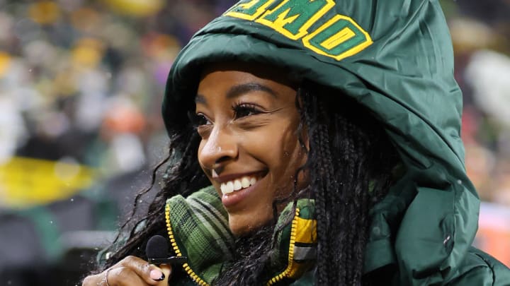 Simone Biles poses in a green jacket with her husband’s name stitched on the hood and smiles to the side.