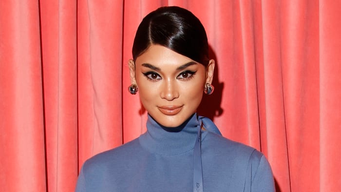 Pia Wurtzbach poses in a blue knit turtleneck dress and a slicked-back updo in front of a neon pink curtain.