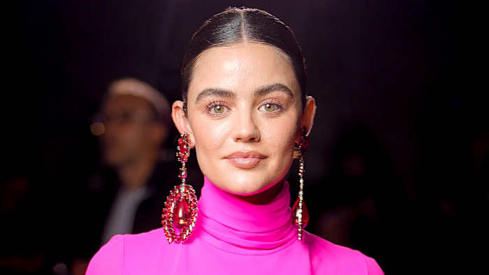 Lucy Hale smiles at the camera wearing a pink turtleneck and oversized red gem earrings.