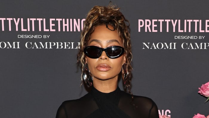 La La Anthony poses in a black sheer high-neck dress and black sunglasses in front of a gray backdrop.