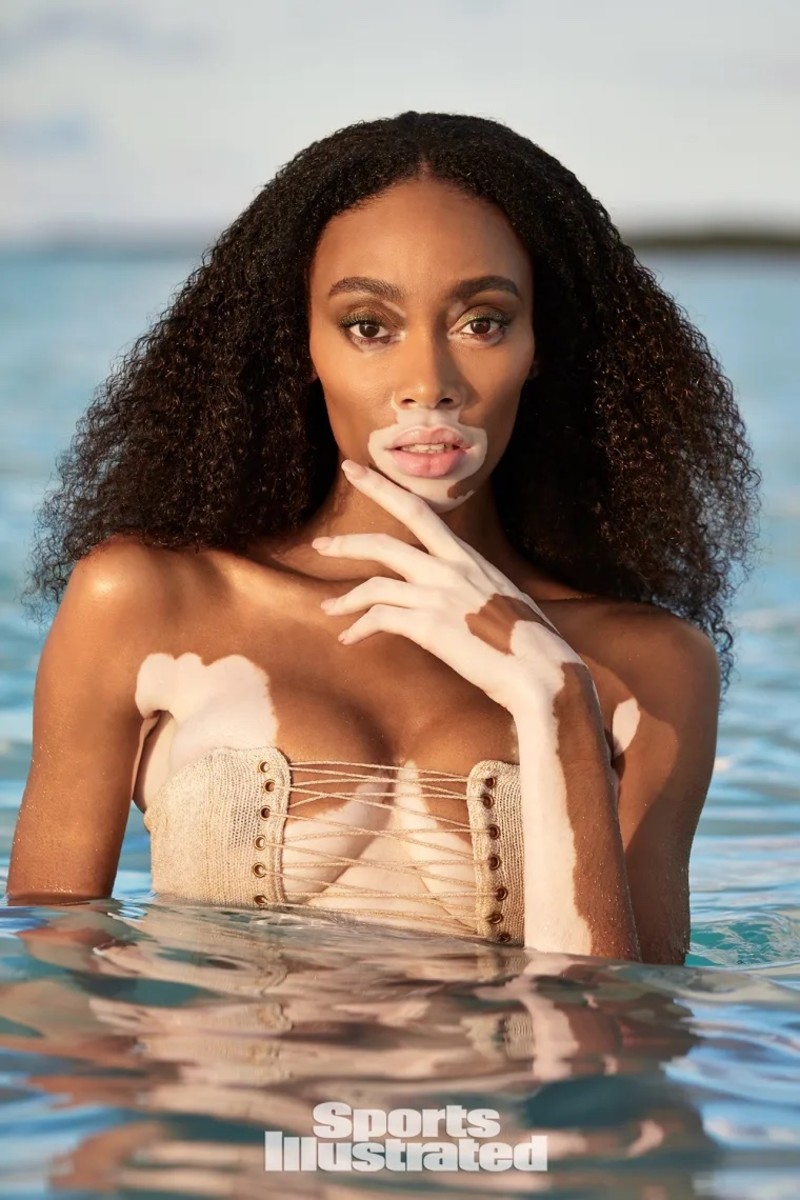 Winnie Harlow sits in the ocean and looks into the camera, wearing a knit bikini top.