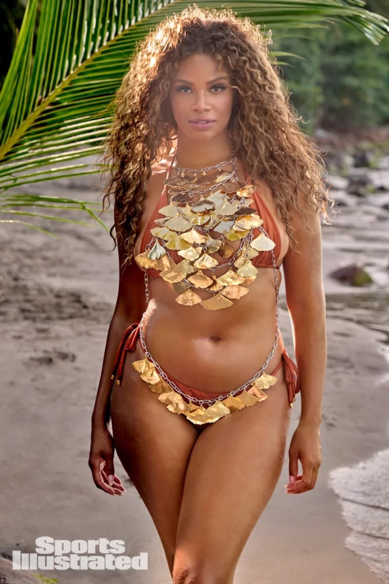 Kame Crawford poses in front of a palm frond in a burnt orange bikini and gold body chain.