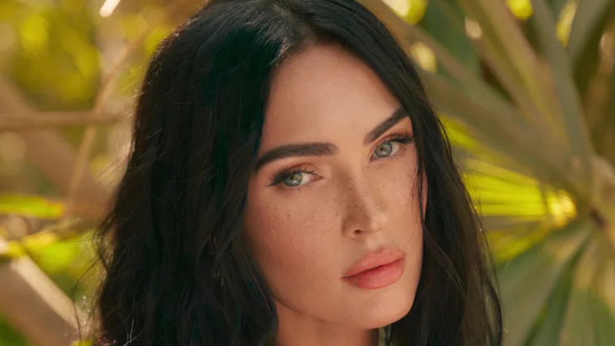 Megan Fox poses for the camera in front of vibrant green palm trees.