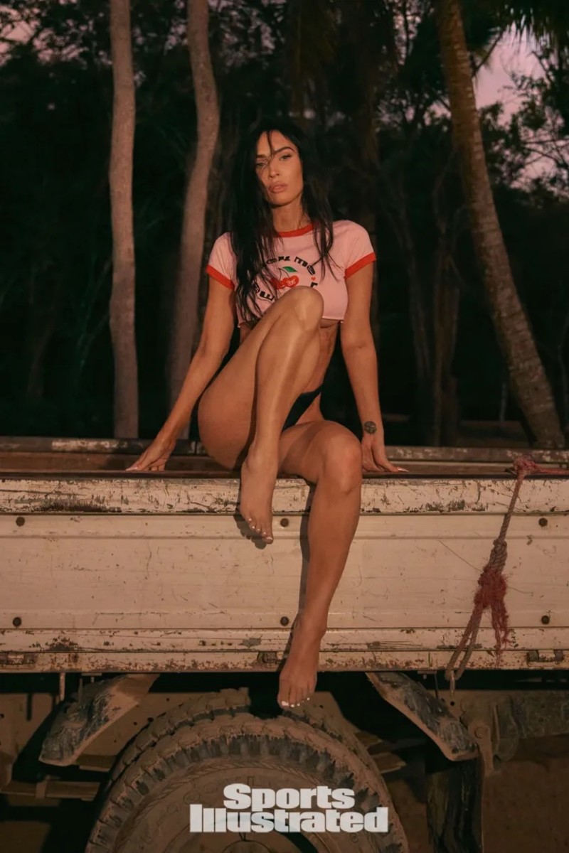 Megan Fox poses in a baby cropped tee and black high-waisted bottoms in front of palm trees.