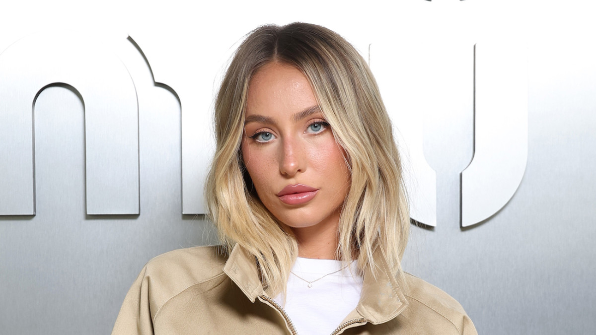 Alix Earle poses in front of a silver backdrop wearing a white tee and tan jacket and her blonde hair down.