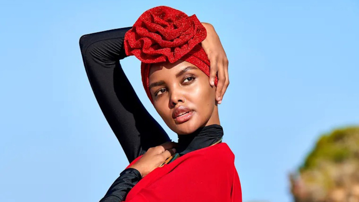 Halima Aden poses in a black catsuit, red kimono, and red rose turban with her arm above her head.