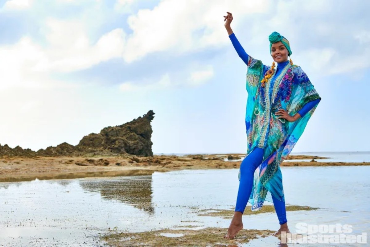 Halima Aden poses in the shallow water with her arm on her hip, wearing a blue catsuit and colorful kimono.