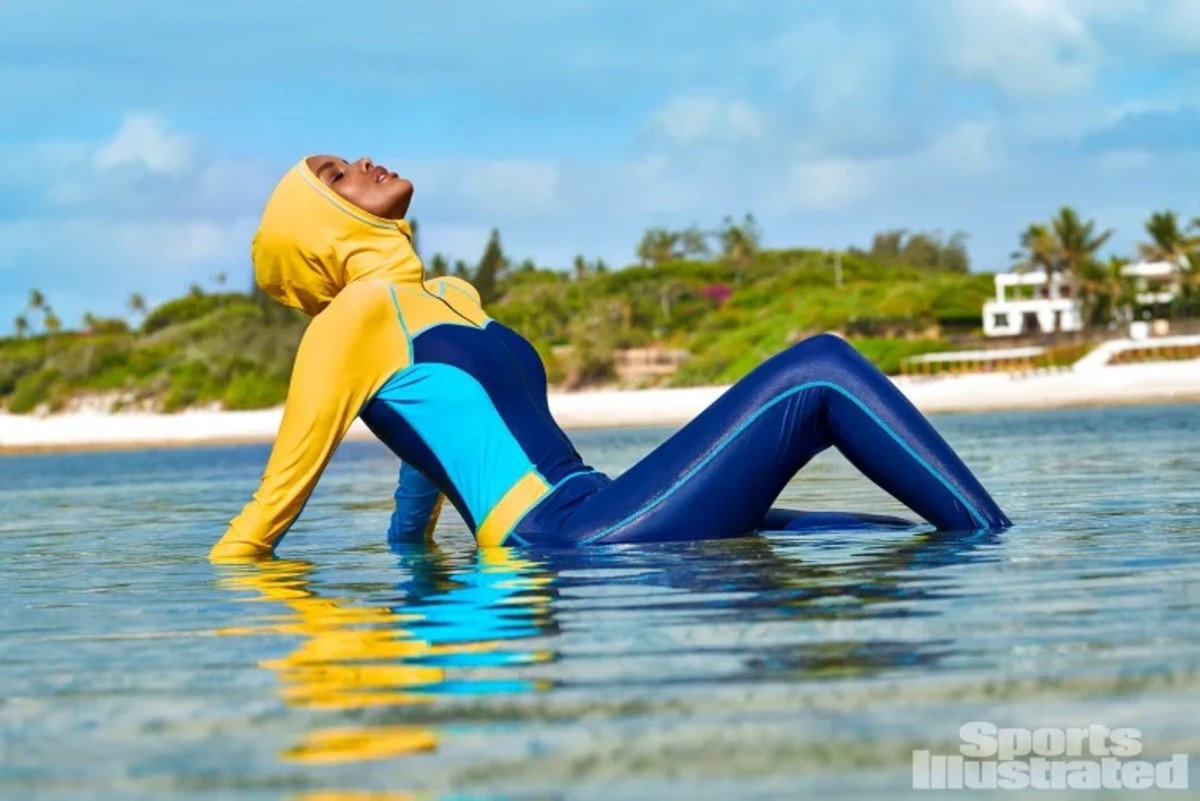 Halima Aden sits in the shallow water wearing a navy, light blue and yellow catsuit.