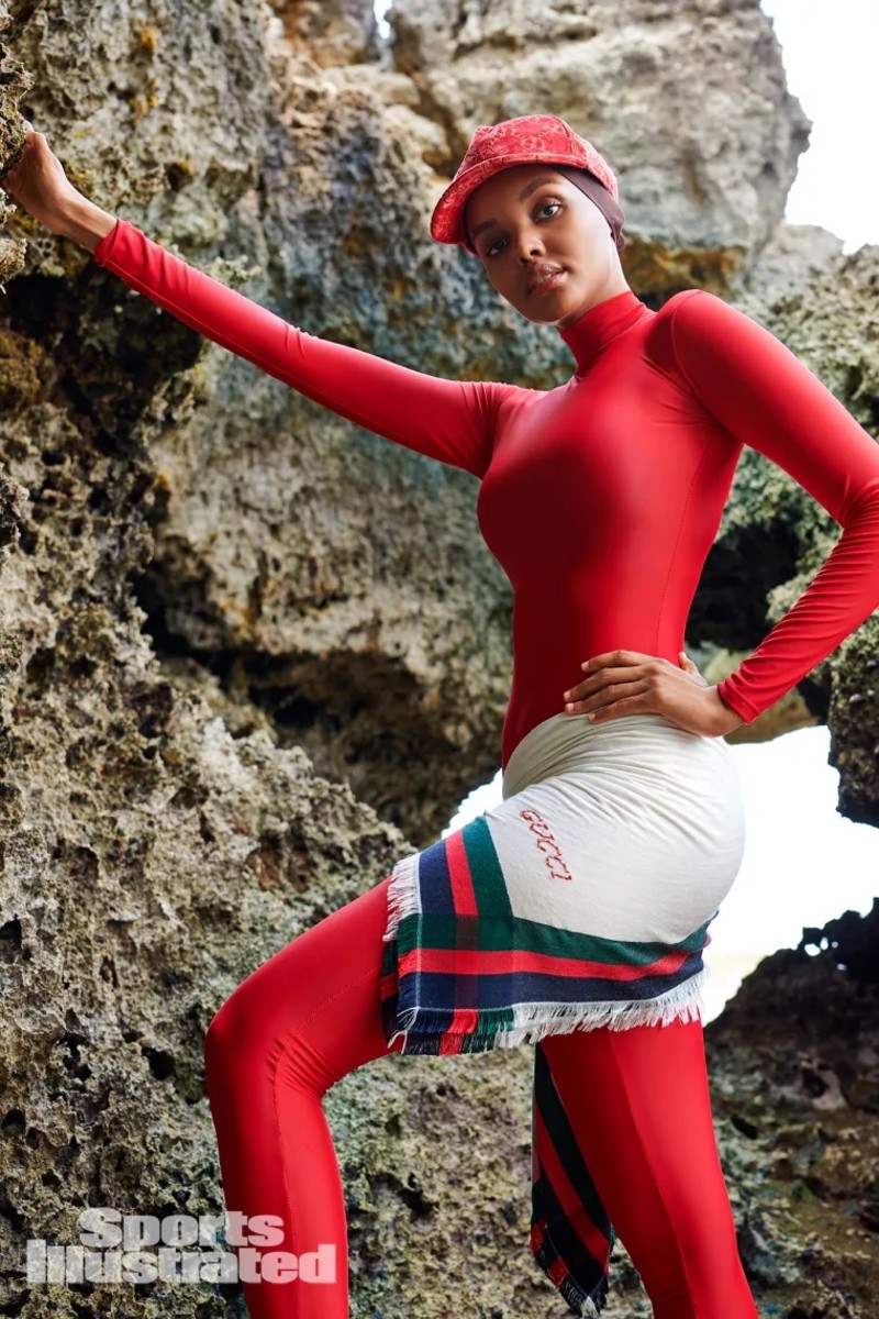 Halima Aden wears a red catsuit, a red hat and a Gucci scarf around her waist and poses with her hand resting on the natural rock beside her.