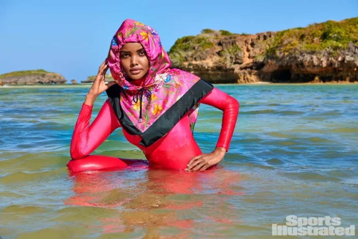 Halima Aden sits in the ocean in a pink catsuit and pink floral headscarf.