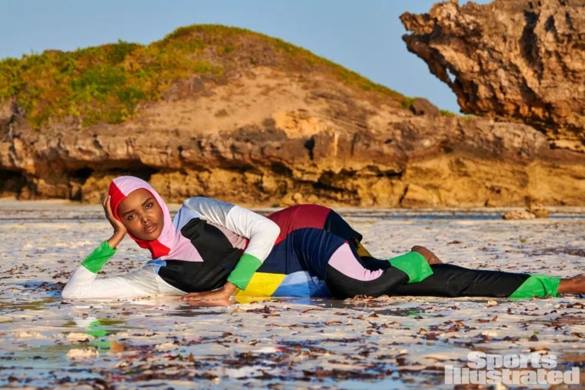 Halima Aden wears a color block catsuit and lays on the ground in front of natural rock formations.