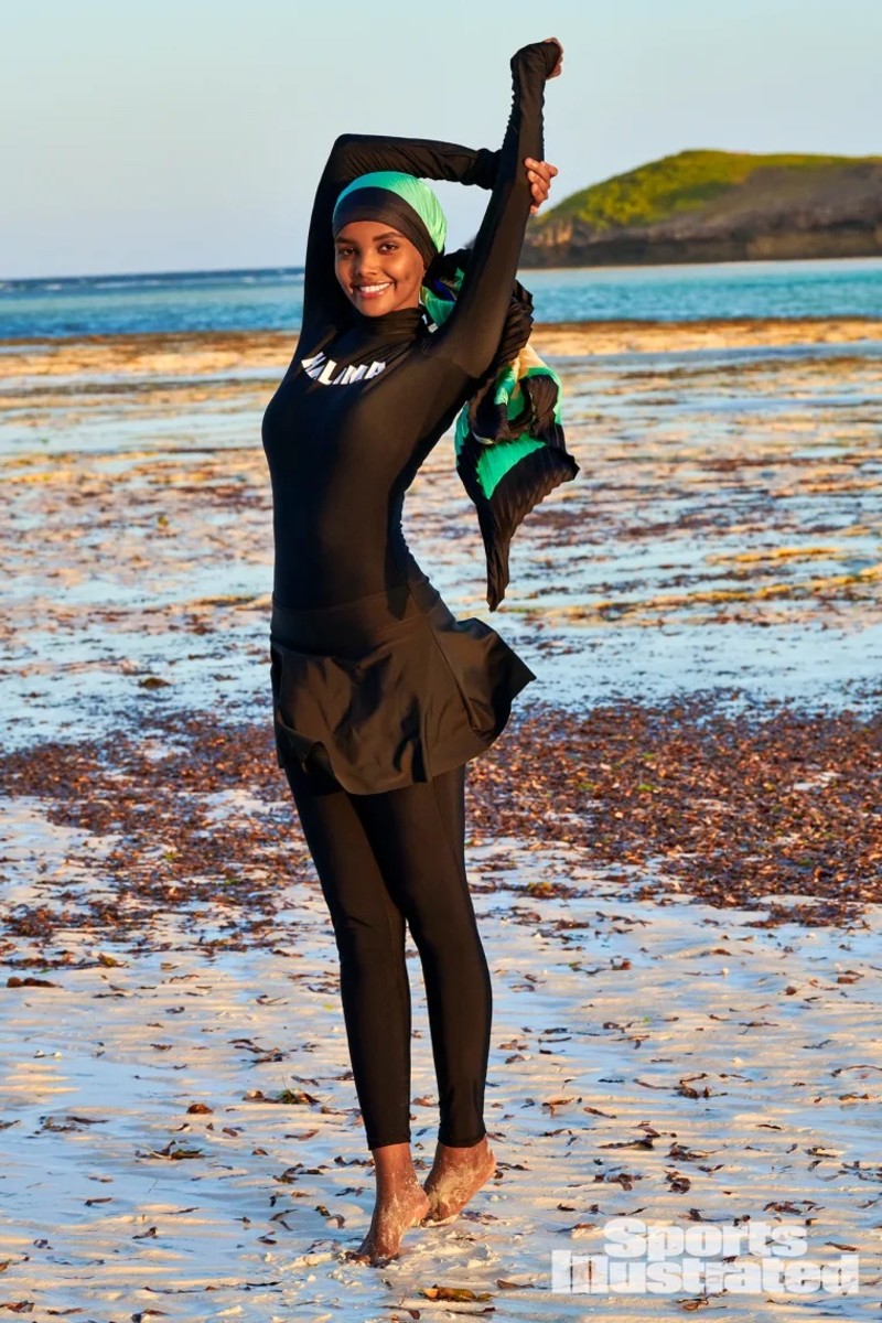 Halima Aden wears a black catsuit and a green and black headscarf and poses with her arms above her head.