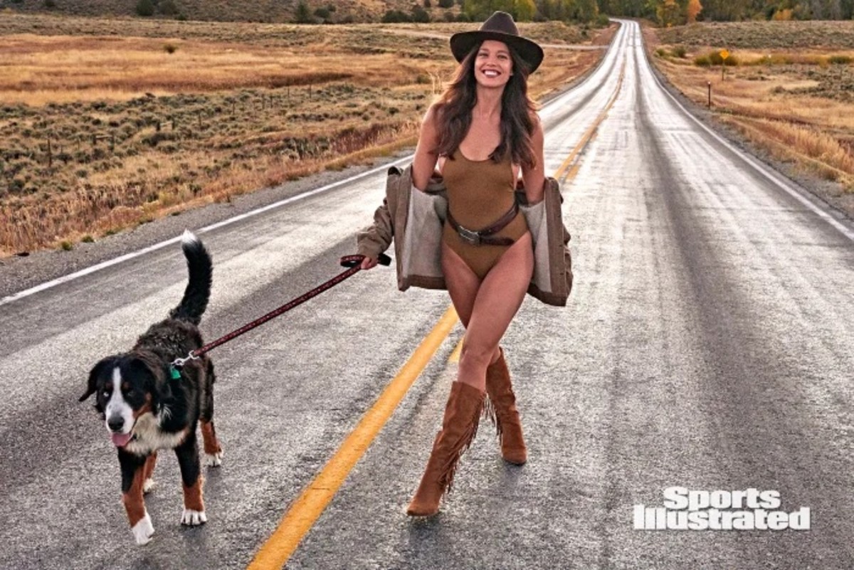Emily DiDonato walks down a Wyoming road wearing a brown one-piece, jacket, fringe boots and a brown hat. She holds the leash of the black, brown and white dog walking beside her.