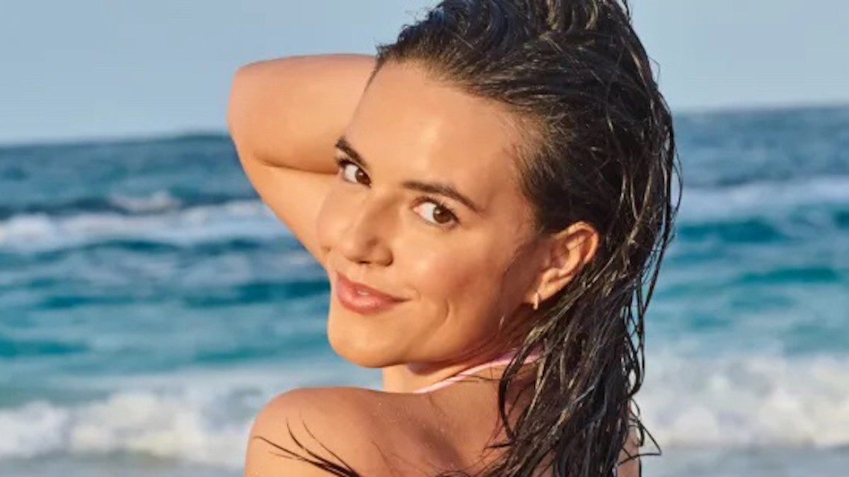 Mady Dewey stands in front of the blue ocean and smiles over her shoulder.