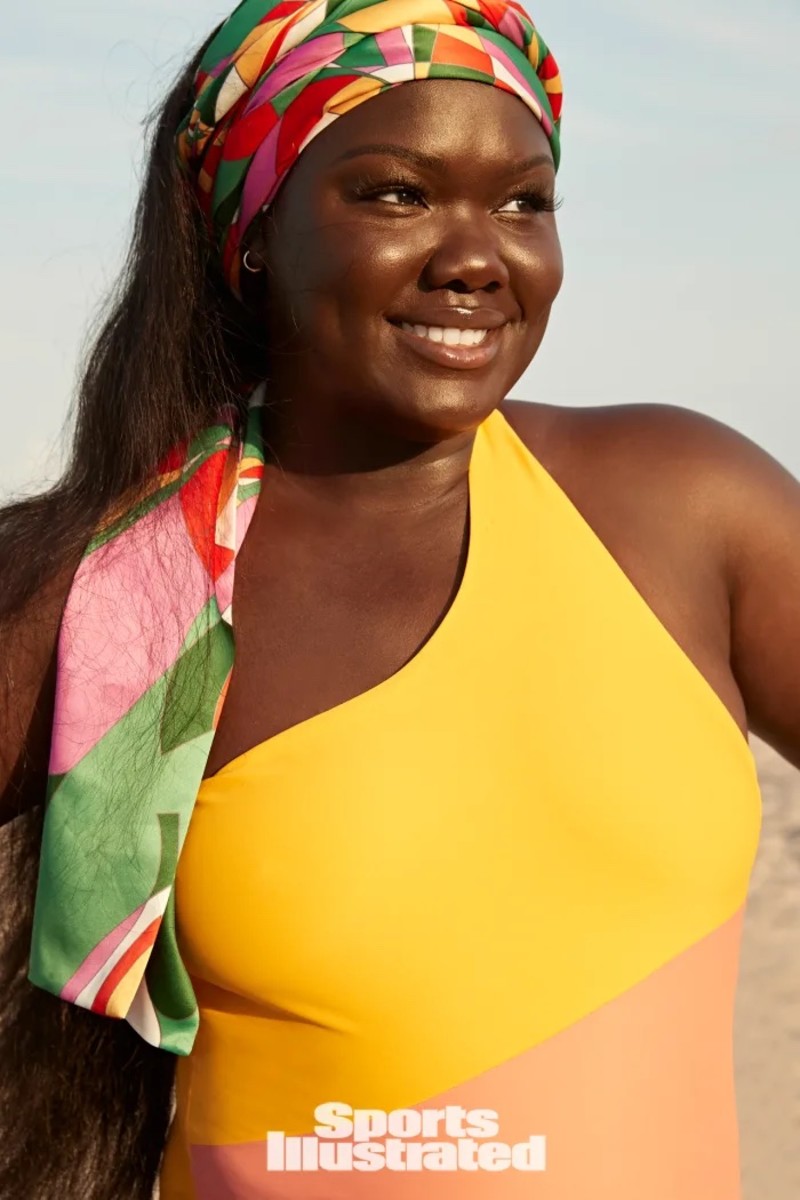 Nyma Tang poses on the beach in a yellow and salmon color block one-shouldered one-piece and a vibrant multicolored headscarf.