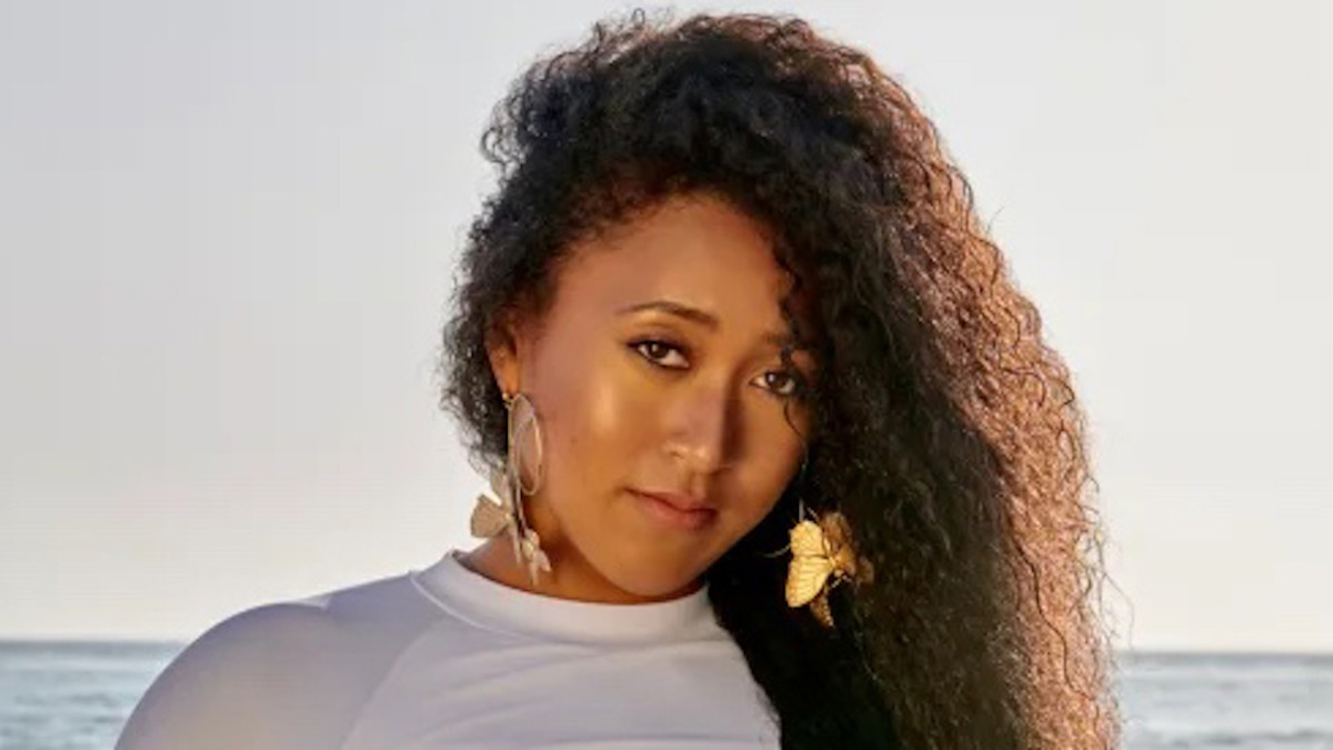 Naomi Osaka stands before the ocean in a white cropped tee and her curly hair parted to one side.