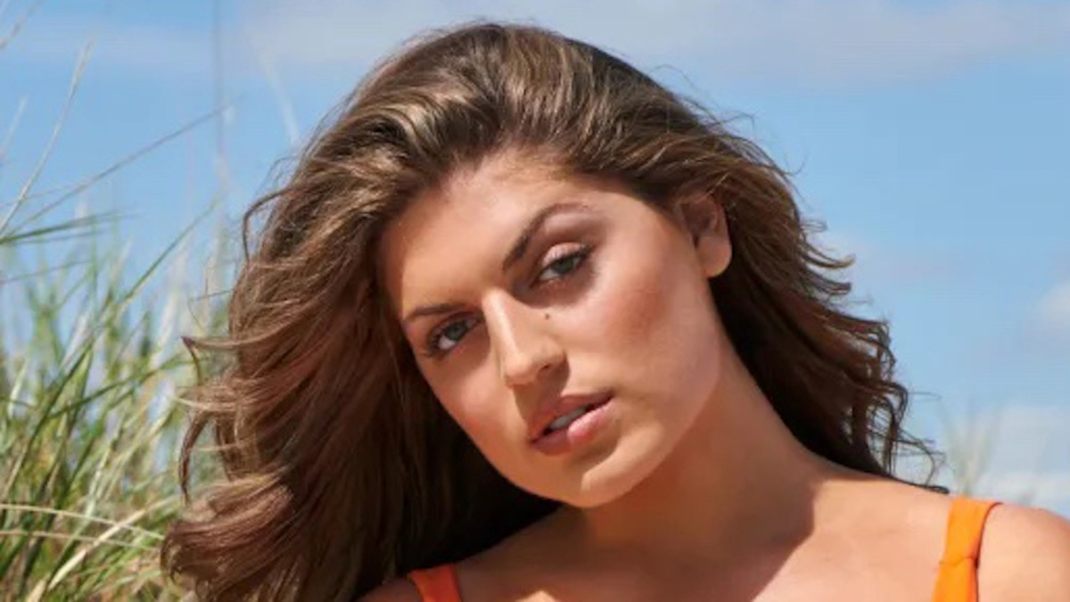 Gabriella Halikas poses in front of the sand dunes in an orange bikini.