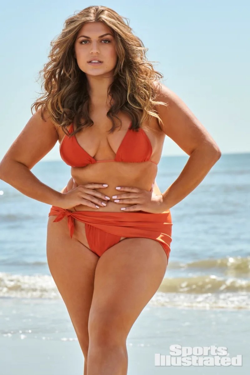 Gabriella Halikas stands in front of the ocean in a deep orange triangle bikini and bottoms.