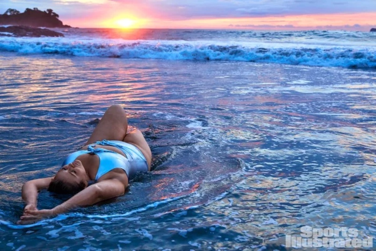 Hunter McGrady lays in the shallow water before the pink and orange sunset in a metallic silver one-piece.