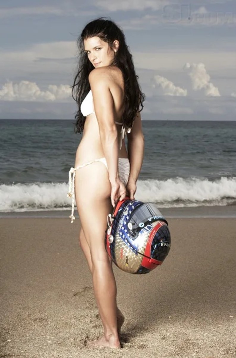 Danica Patrick poses in front of the ocean in a white bikini and holds her race car helmet behind her.