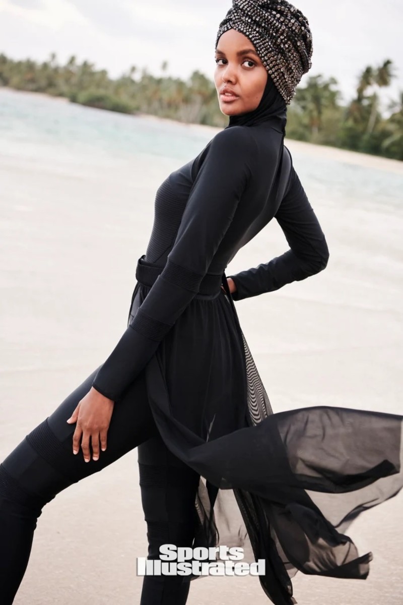 Halima Aden poses in a black burkini and black hijab in front of the ocean.