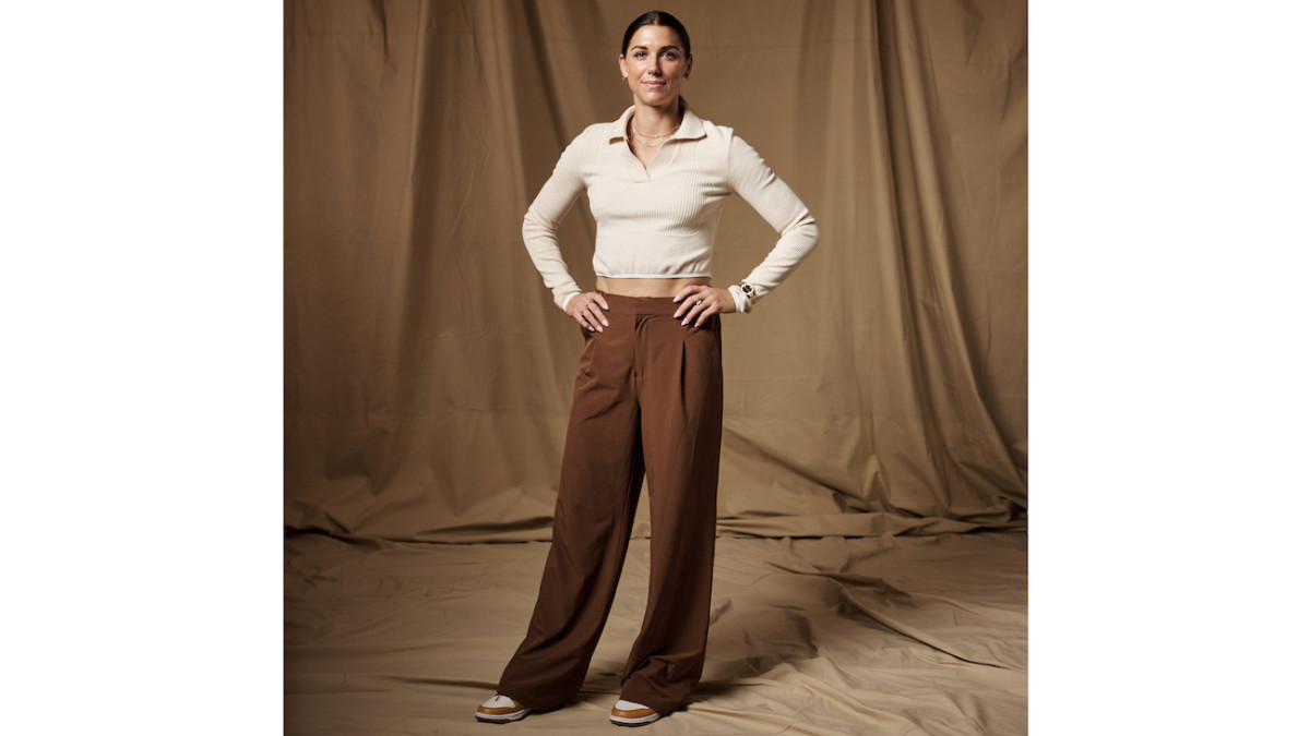 Alex Morgan poses in brown pleated pants and a long-sleeve cream-colored top.