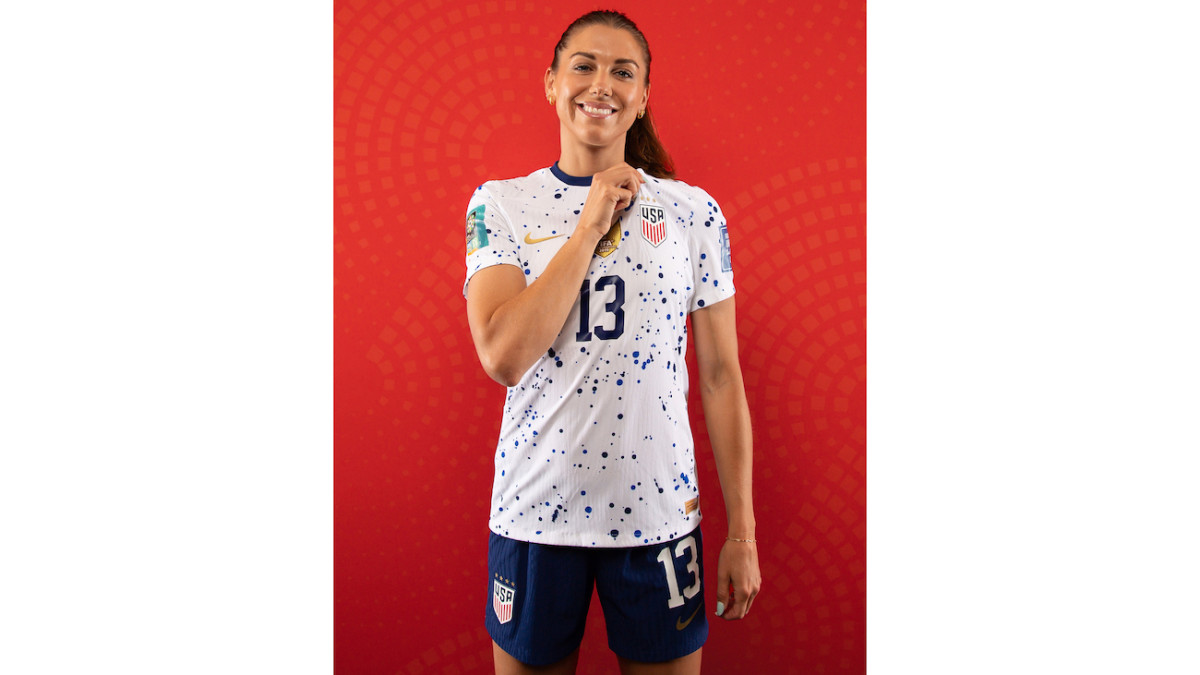 Alex Morgan poses in her USWNT kit in front of a red backdrop.