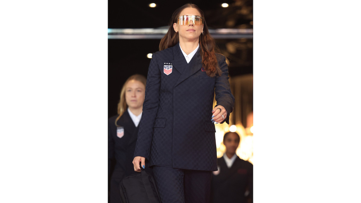 Alex Morgan walks out in a patterned navy suit and light tinted sunglasses.