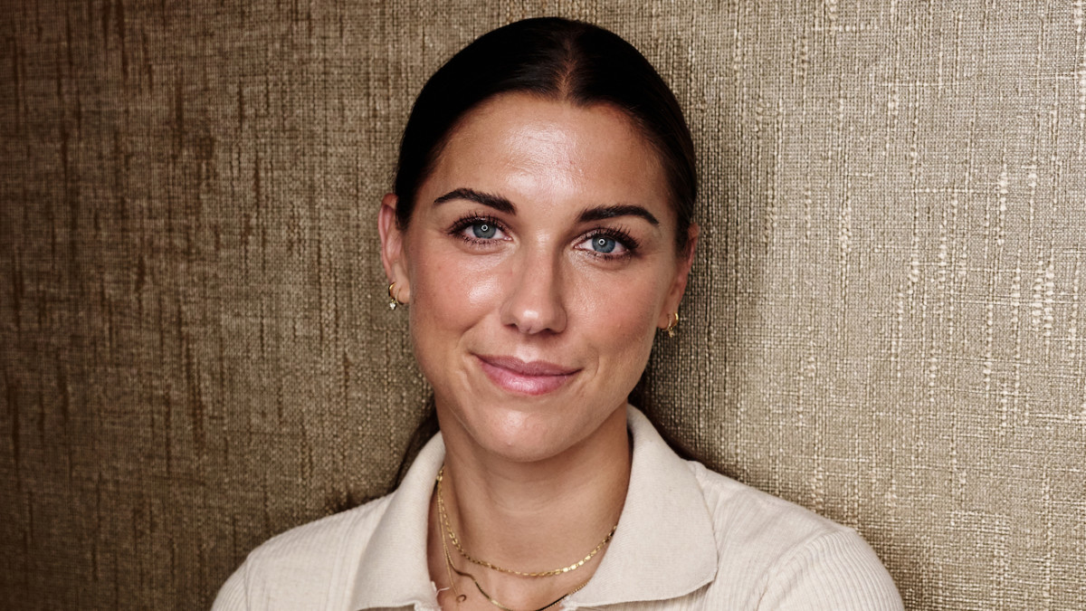 Alex Morgan poses before a textured gold backdrop in a cream-colored sweater and gold necklaces.