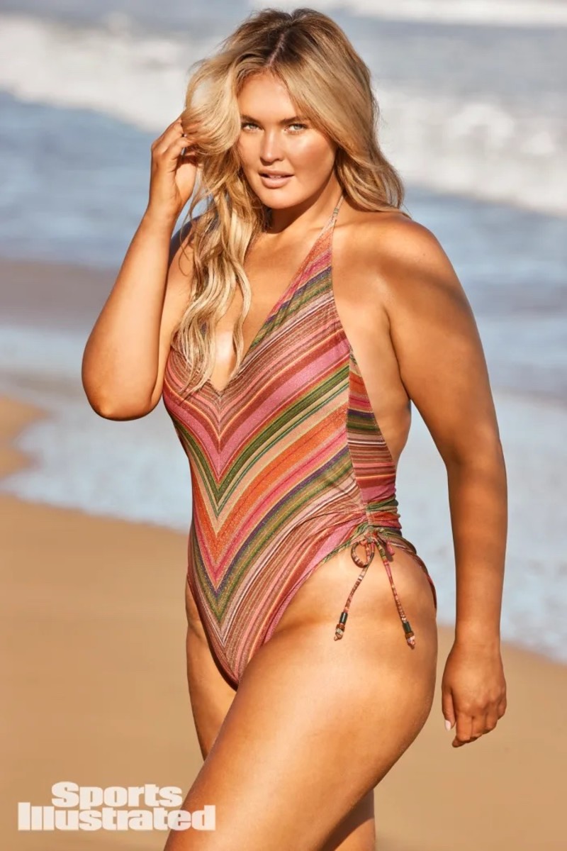 Georgina Burke stands on the beach in a plunging striped green and pink swimsuit.